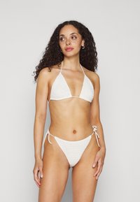 Biquíni branco de croché com top em triângulo e parte de baixo amarrada dos lados. O material tem uma textura padrão, criando um visual de roupa de praia.