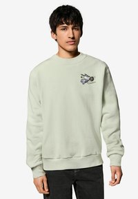 Mintgrüner Pullover aus weichem Stoff. Mit einem kleinen bestickten Cartoon-Charakter, der Tennis spielt, auf der Brust. Klassisches Rundhals-Design.