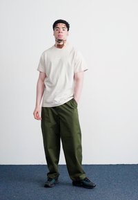 Ανοιχτό μπεζ μπλουζάκι με μικρό λογότυπο, συνδυασμένο με φαρδιά σκούρα πράσινα παντελόνια και μαύρα αθλητικά παπούτσια, σε ουδέτερο φόντο.