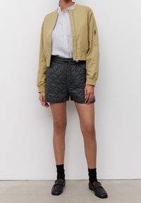 Giacca bomber khaki con tasca con zip sulla manica, abbinata a pantaloni corti grigi trapuntati, calze nere e mocassini neri su un pavimento chiaro.