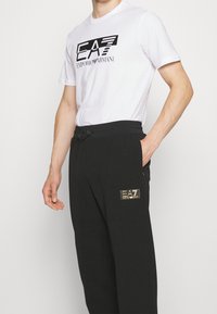 Vit t-shirt med kort ärm och svart logotryck, i kombination med svarta byxor med textilstruktur och en guldlogolapp på fickan.