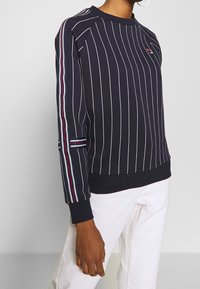 Sudadera azul marino con rayas verticales blancas, puños de canalé y un logo en el pecho. Presenta detalles coloridos en las mangas.