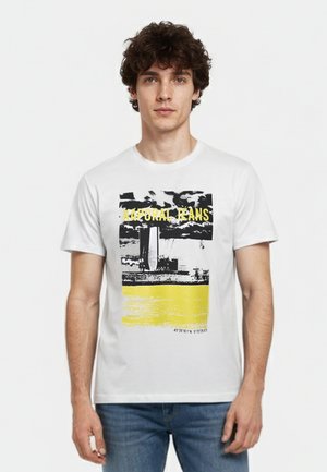 Jonge man met krullend haar die een wit T-shirt draagt met een zwart-gele grafische wolkenkrabber en de tekst "Kaporal Jeans."