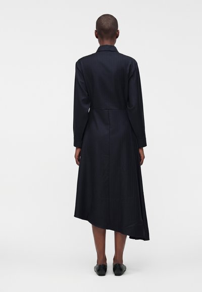 Personne portant une robe midi asymétrique noire à manches longues avec un col et des chaussures plates noires, vue de dos sur un fond uni.