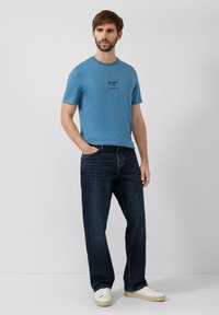 Jasnoniebieska koszulka z krótkim rękawem z nadrukiem na przodzie, zestawiona z ciemnoniebieskimi prostymi jeansami i białymi trampkami.