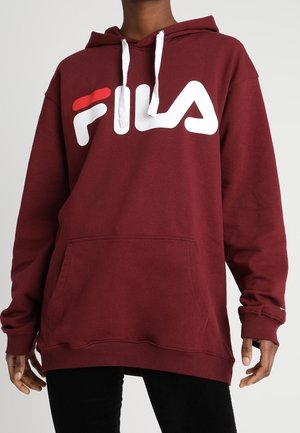 Osoba w bordowej bluzie z kapturem FILA z białymi sznurkami i kieszenią z przodu, ubrana w czarne spodnie na jednolitym tle.