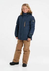 Giacca puffer blu navy con tasca sul petto con zip, pants impermeabili marroni e stivali neri; presenta un cappuccio e una trama trapuntata.