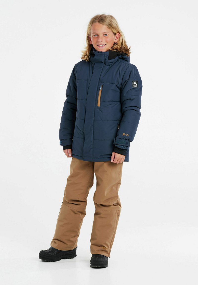 Giacca puffer blu navy con tasca sul petto con zip, pants impermeabili marroni e stivali neri; presenta un cappuccio e una trama trapuntata.