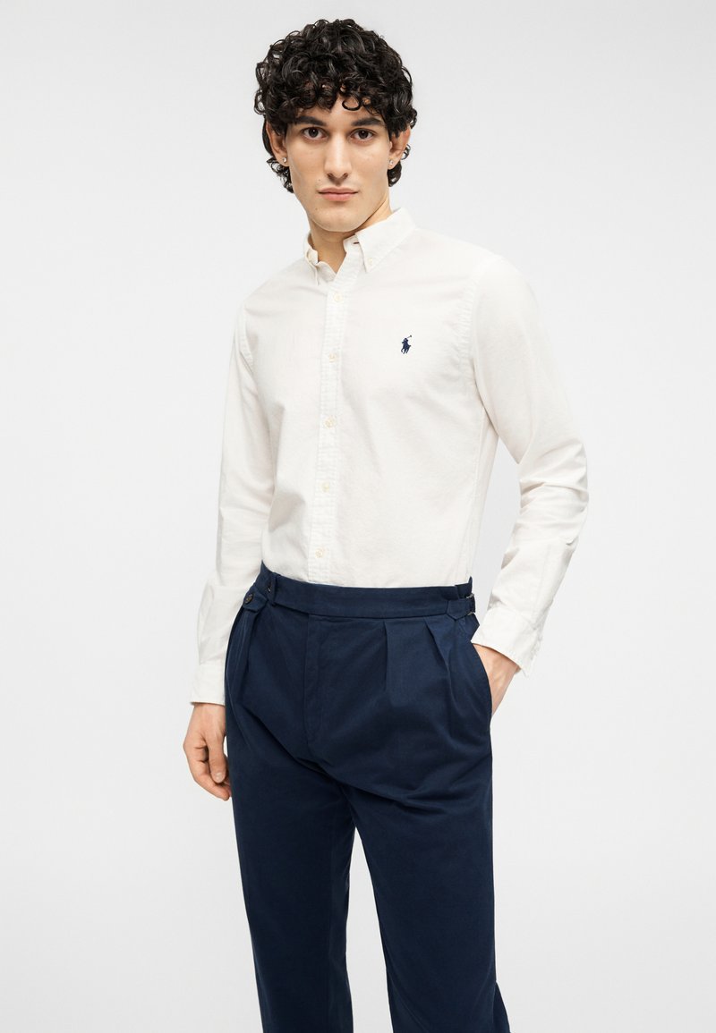 Chemise blanche boutonnée à manches longues avec un col classique, ornée d'un petit logo bleu. Associée à un pantalon plissé bleu marine.