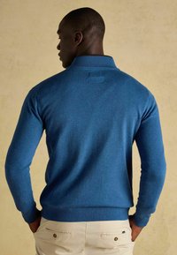Zils adīts džemperis ar augstu apkakli, ribotu aproci un apakšu. Aizmugures skats atklāj gludu tekstūru un piegulošu dizainu.