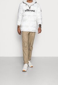 Uomo che indossa un giubbotto imbottito Ellesse bianco, pantaloni beige arrotolati e sneakers bianche, in piedi contro uno sfondo bianco uniforme.