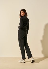 Combinaison en denim noir avec une taille cintrée et des manches longues, dotée de coutures marquées et de poches latérales, associée à des baskets blanches.