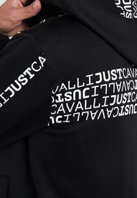 Czarny sweatshirt z wyraźnym białym napisem w różnych orientacjach. Projekt zajmuje obszary ramion i rękawów, z gładką teksturą.