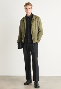 Oliven grøn bomberjakke med lynlås foran, sort skjorte med knapper, sorte skræddersyede bukser og sorte støvler. Minimalistisk design, teksturerede materialer.
