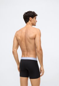 Svarta bomullsboxershorts med ett ljusgrått elastiskt midjeband med texten "Calvin Klein". Modellen visas bakifrån.