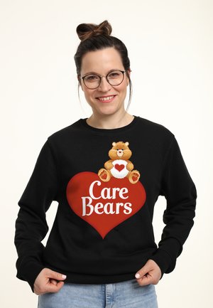 Donna sorridente con occhiali che indossa una felpa nera con il logo dei Care Bears, raffigurante un orsacchiotto che tiene un cuore sopra una forma a cuore rossa.