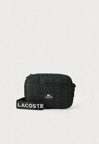 Tumši zaļa taisnstūra soma ar ģeometrisku rakstu, melnu rāvējslēdzēju, priekšējo kabatu un siksnu ar uzrakstu "LACOSTE" baltā krāsā.