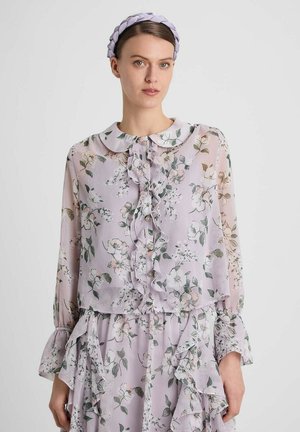 Frau mit geflochtenem Haarreif trägt eine hellviolette Bluse mit Blumenmuster und Rüschen vorne sowie einen passenden Rock vor einem schlichten Hintergrund.