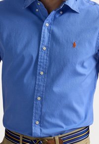 Světlomodrá košile na zapínání s materiálem odolným proti mačkání, zdobená malým oranžovým logem na hrudi a bílými knoflíky. Detaily s vybraným prošitím.