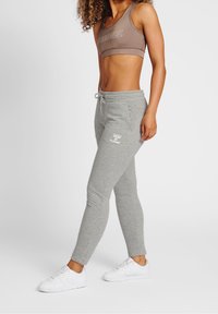 Pantalones de chándal grises con cintura ajustable y logo en el muslo, combinados con un sujetador deportivo marrón y zapatillas blancas. Tejido suave, diseño ajustado.