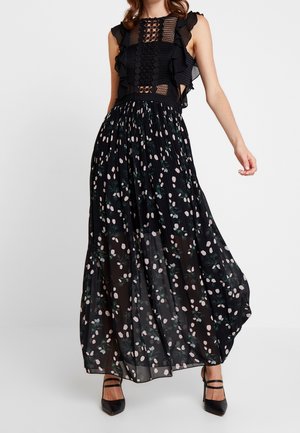 Femme portant une robe maxi noire sans manches avec un corsage en dentelle et à volants et une jupe plissée avec un motif floral rose pâle et vert.
