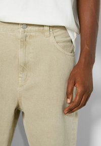 Jeans in denim beige con una finitura testurizzata, dotati di tasche frontali e chiusura con bottone metallico. Mano appoggiata sul lato dei jeans.