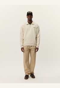 Sudadera cremosa con cremallera y cuello, detalle del logo en el pecho, combinada con pantalones beige y zapatos marrones de cordones. Gorra negra con logo.