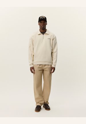 Sweatshirt crème avec zip et col, détail de logo sur la poitrine, associé à un pantalon beige et des chaussures marron à lacets. Casquette noire avec logo.