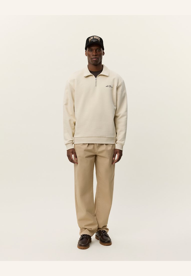 Sudadera cremosa con cremallera y cuello, detalle del logo en el pecho, combinada con pantalones beige y zapatos marrones de cordones. Gorra negra con logo.