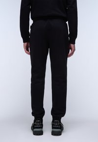 Man staat met zijn rug naar ons toe, gekleed in zwarte joggpants en zwarte sneakers met grijze accenten, op een witte achtergrond.