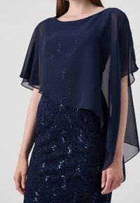 Cape en chiffon bleu marine présentant un design fluide sur une robe ajustée en dentelle bleue marine ornée de paillettes, créant une texture superposée et une profondeur visuelle.
