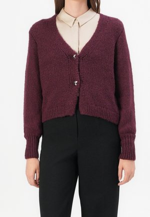 Femme portant un cardigan en maille violet avec deux boutons argentés sur une chemise beige à col et un pantalon noir sur fond blanc.