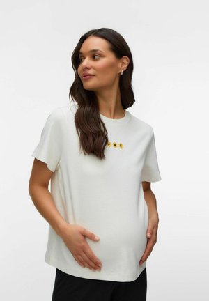 Gravid kvinde med langt mørkt hår iført hvid t-shirt med solsikker, hænderne blidt hvilende på mave, kigger til siden.