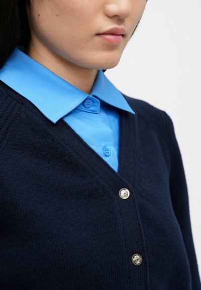 Navy pletený cardigan s V-výstřih a třemi stříbrnými knoflíky, nošený přes světle modrou košili s límečkem. Texturovaná tkanina, vrstvový design.