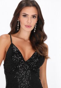 Vestido negro con lentejuelas, escote en V profundo y tirantes finos. Pendientes largos y brillantes complementan el look, realzando el diseño general.
