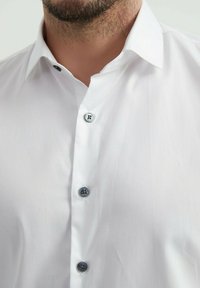 Chemise blanche à col classique, dotée de boutons noirs sur le devant et d'un tissu lisse et texturé.