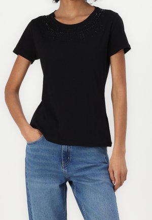 Vrouw met een zwart topje met korte mouwen en subtiele versieringen rond de halslijn, en een blauwe spijkerbroek, één hand in de zak.