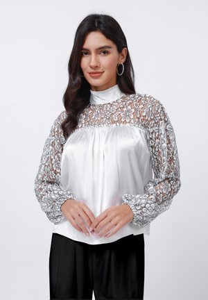 Weißes Blusenshirt mit aufwendigen schwarzen Spitzenlangen Ärmeln, raffinierten Bündchen und hohem Kragen. Glatter Satinoberteil mit dezenten Falten an der Vorderseite.