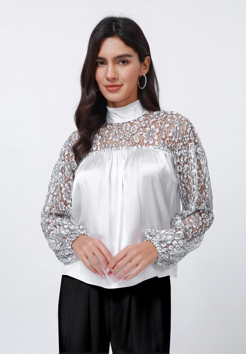 Blusa bianca con intricate maniche lunghe in pizzo nero, polsini arricciati e collo alto. Corpo in satin liscio con leggere pieghe sul davanti.