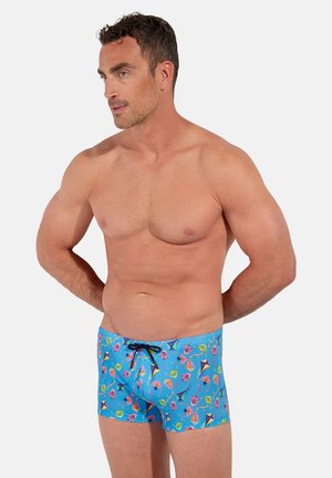 Homme musclé portant un maillot de bain bleu avec des motifs colorés de cerfs-volants, debout les mains derrière le dos, regardant légèrement vers la gauche.