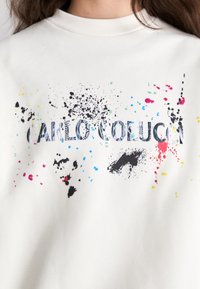 Sweatshirt blanc avec un graphisme texturé et des motifs de gouttes en rose, bleu, vert, et du texte noir indiquant "CARLO COLUCC."