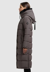 Lange, gewatteerde jas in donker taupe, met een gevoerde capuchon van nepbont, ritszakken en elastische manchetten. Slanke silhouette met een gestructureerde afwerking.