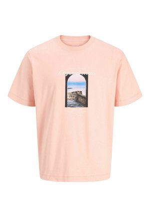 Dreng med krøllet hår iført en lyseblå jakke over en lyserød T-shirt med grafik, cremefarvede shorts, hvide sokker og beige sko, stående mod en hvid baggrund.