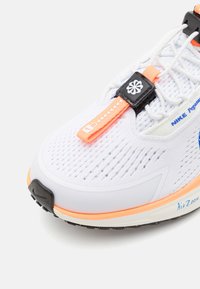 Hvitt sportssko med pustende mesh-oberdel, svarte og oransje detaljer, synlig snøresystem og gummisåle designet for grep.