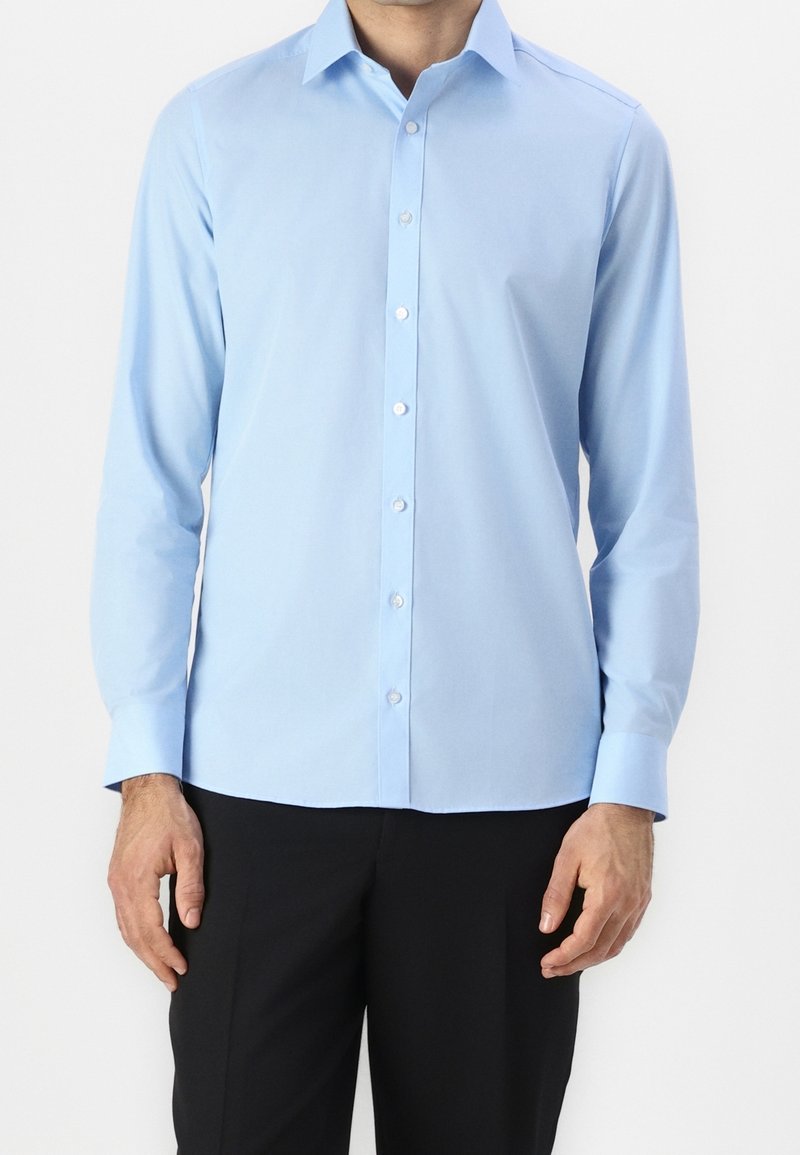 Homme portant une chemise bleu clair à manches longues boutonnée et un pantalon noir, debout devant un arrière-plan uni.