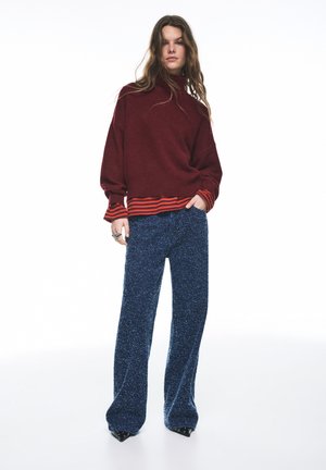 Bordowy sweter z dzianiny z czerwono-pomarańczowymi prążkowanymi mankietami zestawiony z niebieskimi, szerokimi spodniami w cętki oraz czarnymi, szpiczastymi butami.