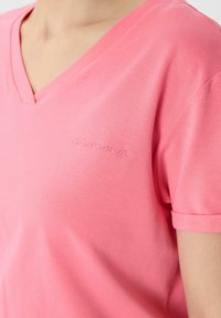 Różowa koszulka z dekoltem w serek, wykonana z gładkiego materiału, z krótkimi rękawami. Subtelne haftowane logo w kształcie przecinka na piersi. Prosty i dopasowany krój.
