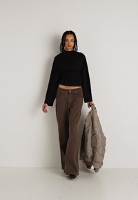 Suéter negro corto, pantalones de pierna ancha marrones y una chaqueta acolchada marrón claro. La modelo sostiene la chaqueta, de pie contra un fondo neutro.