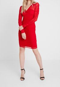 Robe rouge froncée avec manches longues en voile, décolleté en V, longueur au genou et silhouette ajustée. Portée avec des talons noirs à brides.