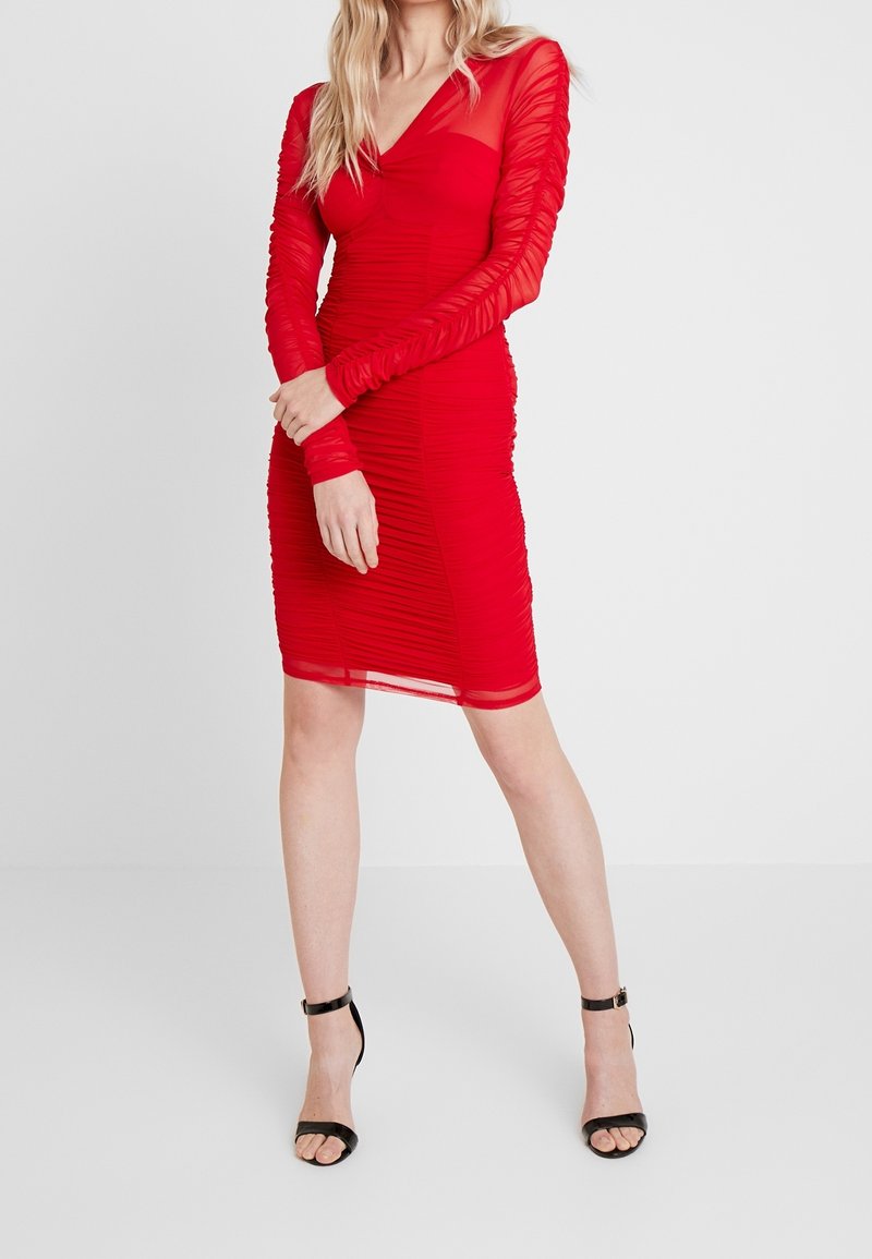 Robe rouge froncée avec manches longues en voile, décolleté en V, longueur au genou et silhouette ajustée. Portée avec des talons noirs à brides.
